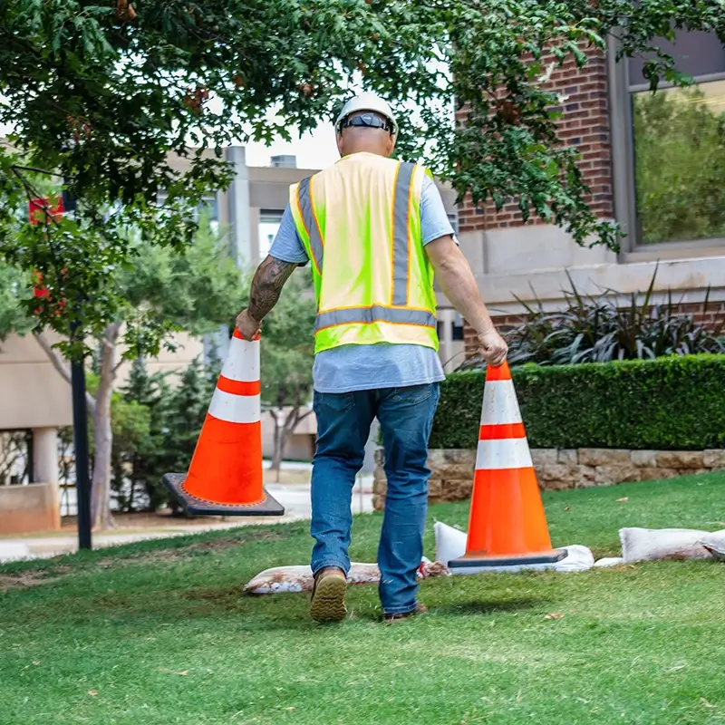 A worker wearing a yellow safety vest carrying two orange cones